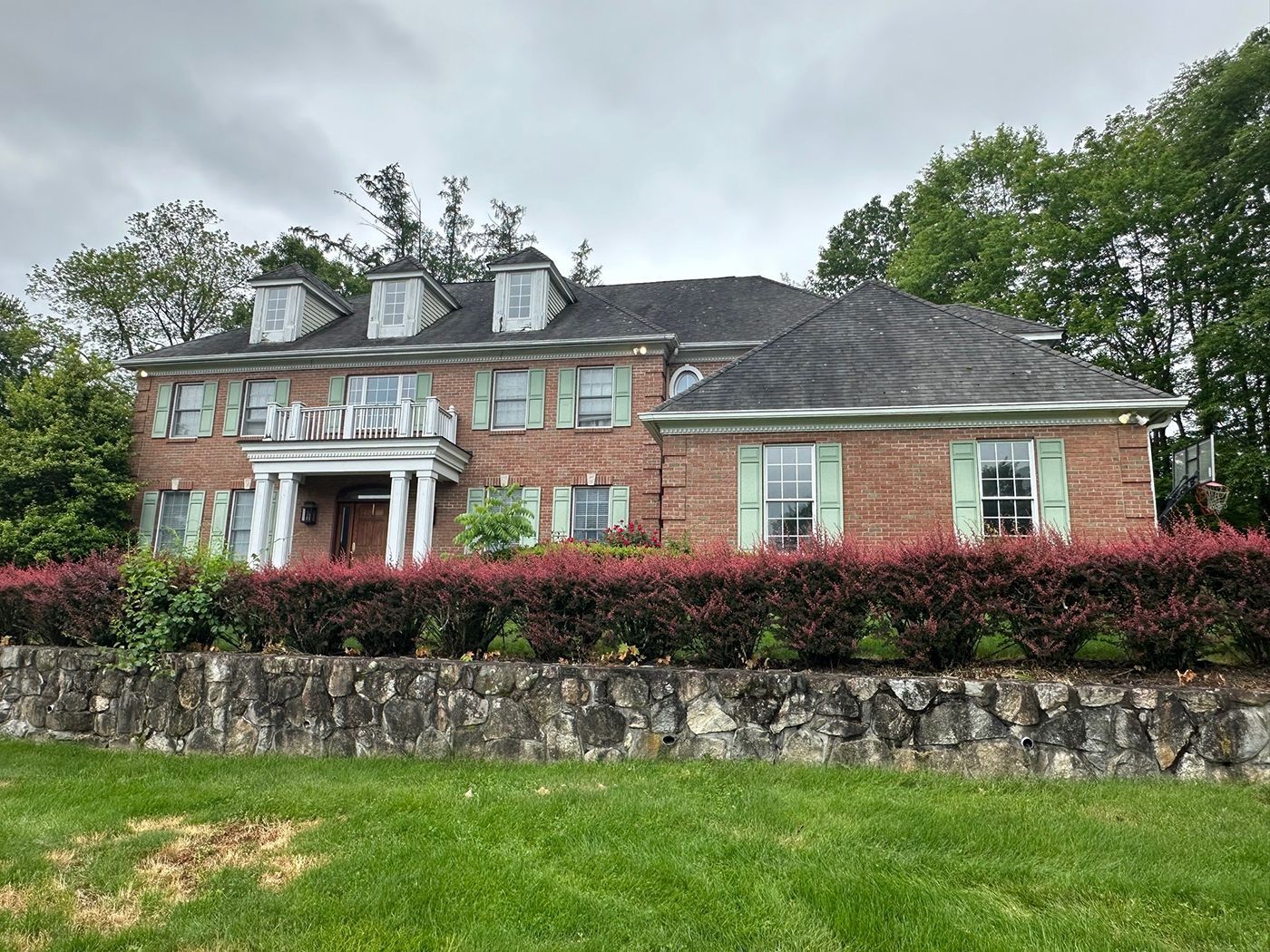 Brick house with white columns, green shutters, and a dark roof sits behind a stone wall 
