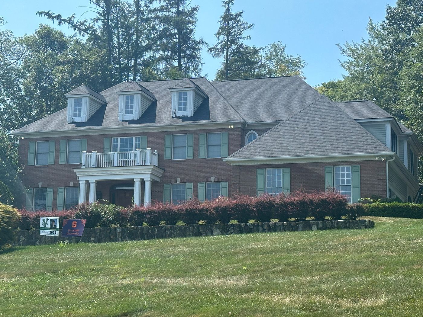 A brick two-story house with a dark gray roof, white pillars, and green shutters, surrounded by trees and a green lawn.