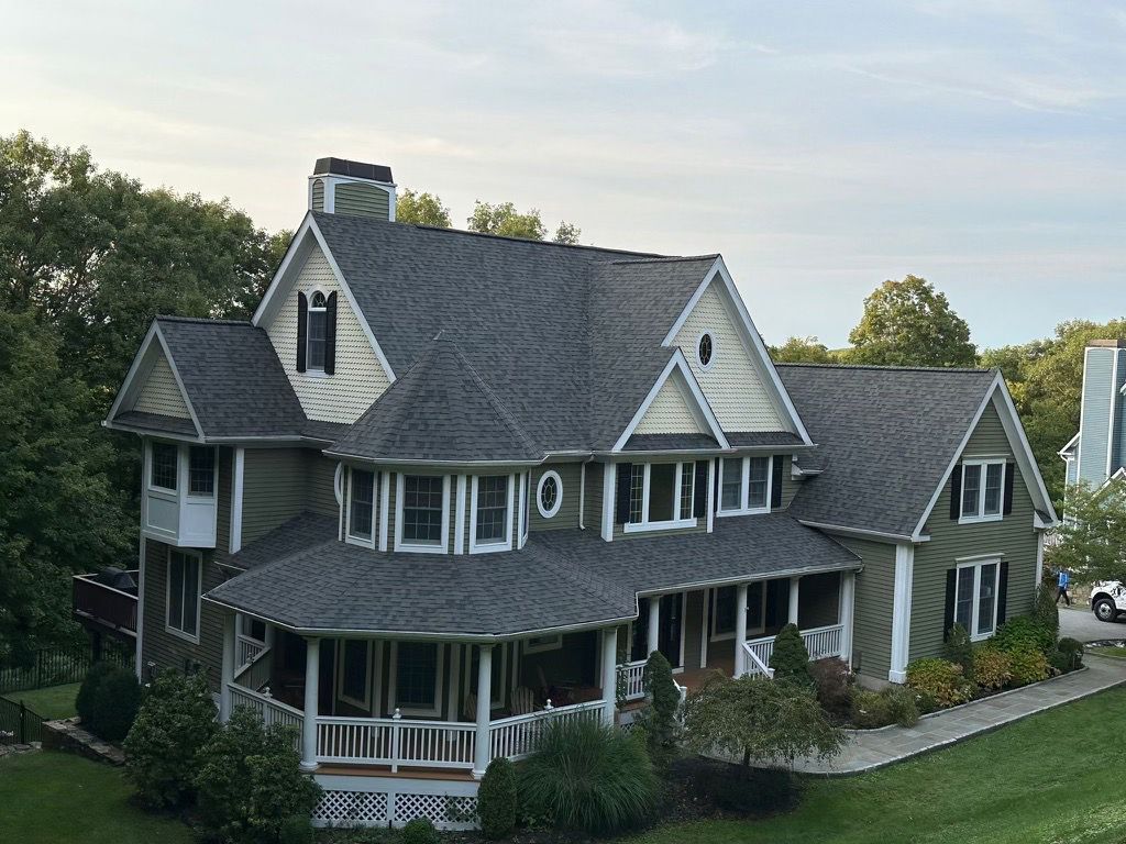 A two-story house with gray shingles and green siding, featuring a wraparound porch and a circular turret