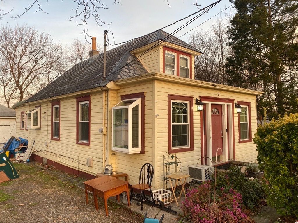 Yellow cottage with red trim, two-story with dormer, small front yard with outdoor furniture, sky in the background.