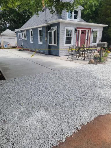 A house with blue siding and a red door has a newly poured concrete driveway, with gravel