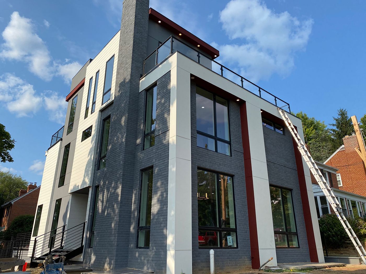 Modern three-story house with gray and white exterior, large windows, and a rooftop deck under a blue sky.