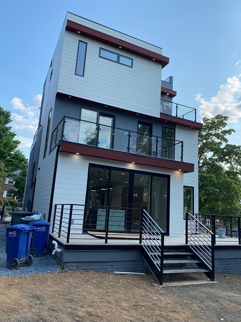 Modern three-story house with white brick facade, glass balconies, and a deck. Blue trash bins sit near the house on gravel.