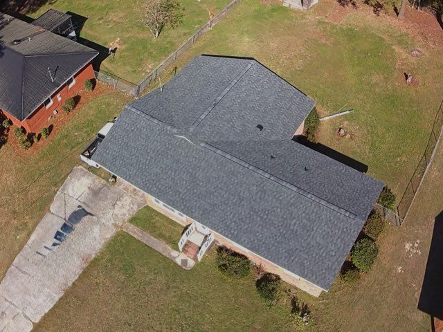 An aerial view of a single-story house with a grey shingled roof, surrounded by a lawn and a concrete driveway.