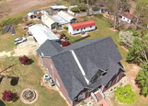 An aerial view of a brick house with a dark roof, adjacent to a smaller outbuilding, a white shed, and a grassy yard.