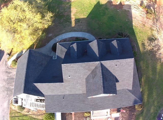 Aerial view of a residential house with a dark gray shingled roof, surrounded by green grass and a paved walkway.