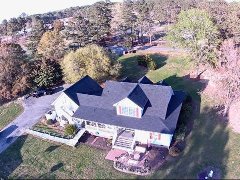 Aerial view of a white two-story house with a dark roof and porch, situated on a grassy, tree-lined lot with a driveway.