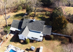 Overhead view of a white house with a dark gray roof, surrounded by trees and grass. A pool is in the foreground.