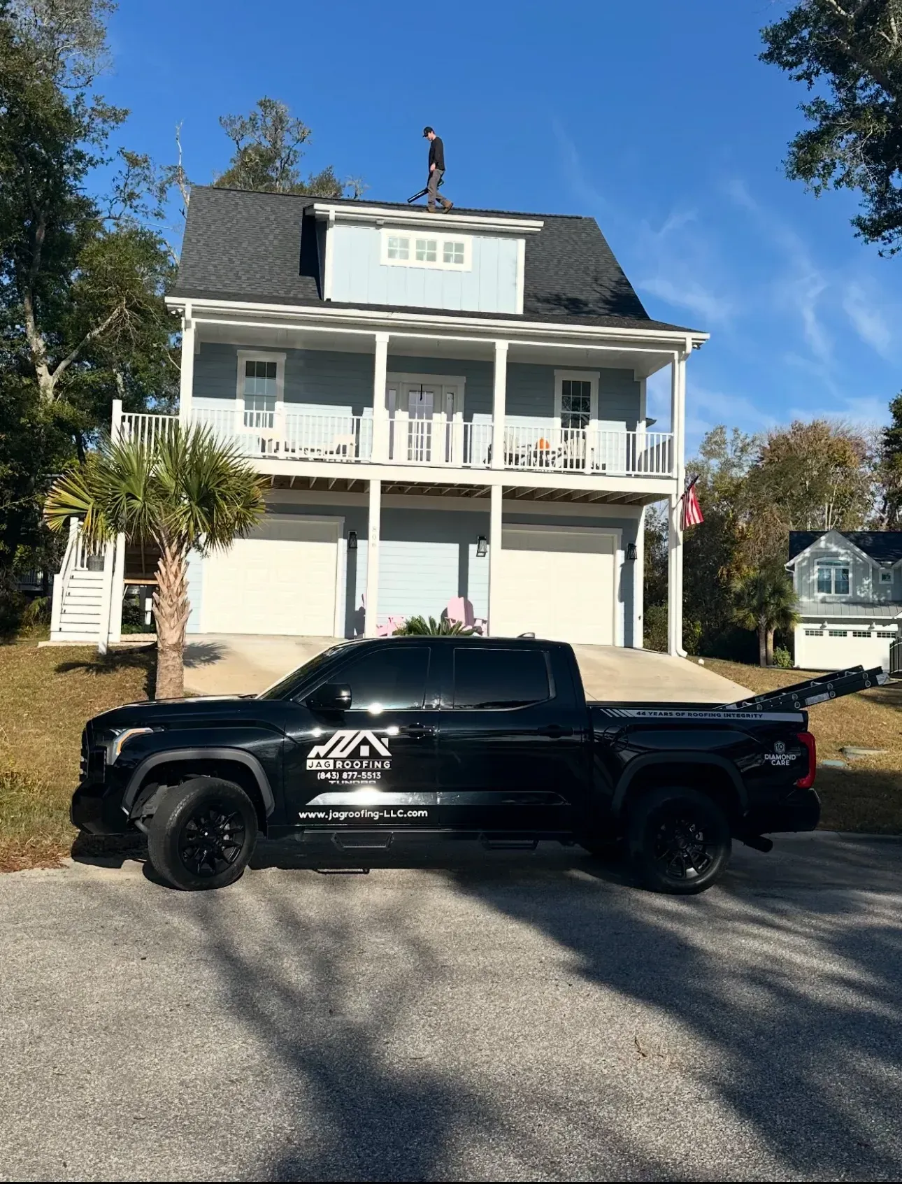 Black truck parked in front of a blue house with a person on the roof. Sunny day.