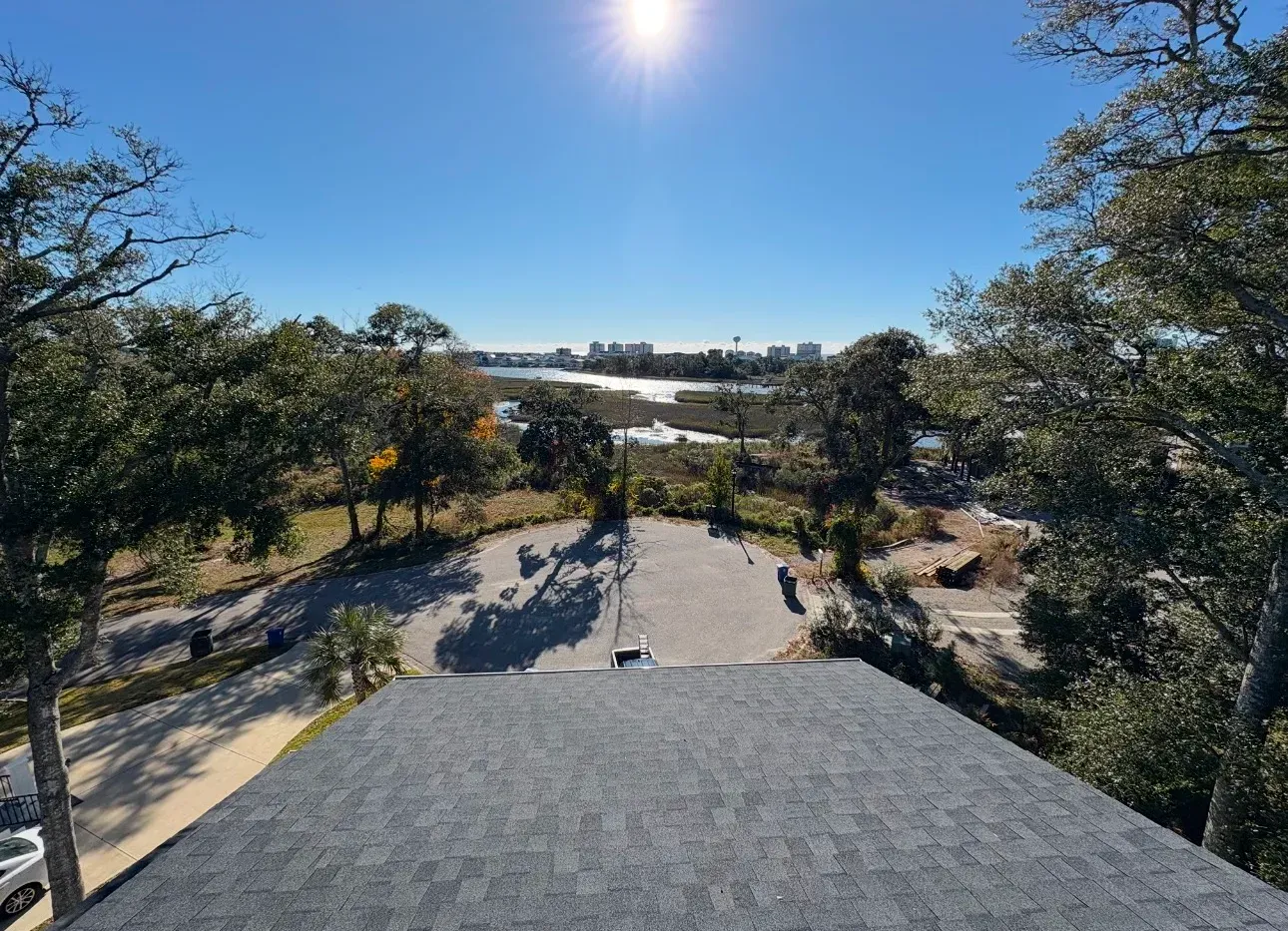 View from above a gray rooftop, looking towards a sunny horizon with trees and water.