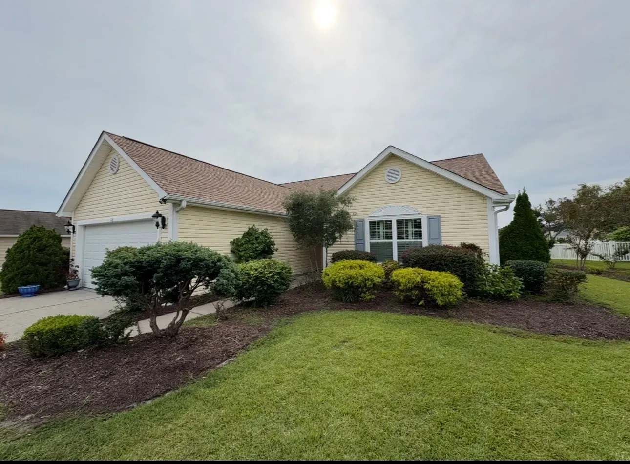 Yellow house with a brown roof, green lawn, and landscaped garden under a cloudy sky.