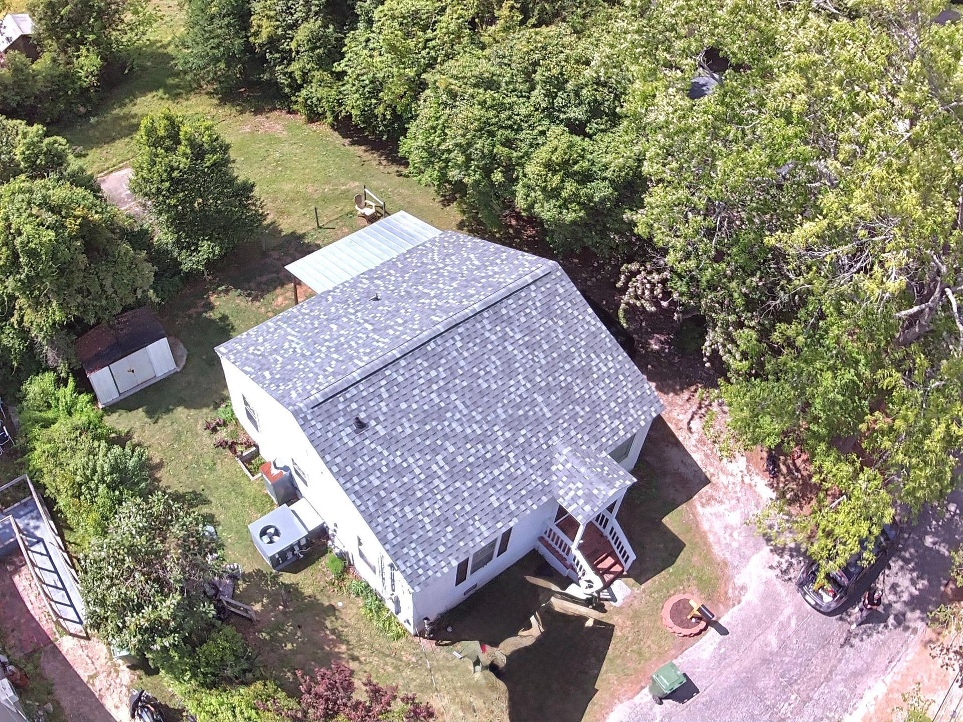An aerial view of a white house with a grey shingled roof, surrounded by trees, a lawn, and a driveway.