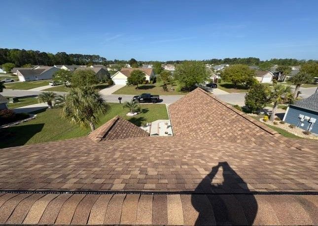 A high-angle view looking down over a brown shingled roof towards a suburban neighborhood with houses and trees.