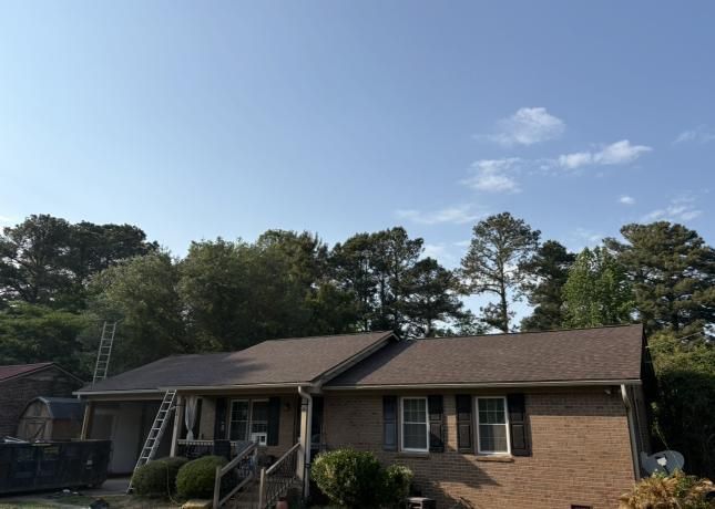 Single-story brick house with brown roof and front porch, framed by trees under a blue sky