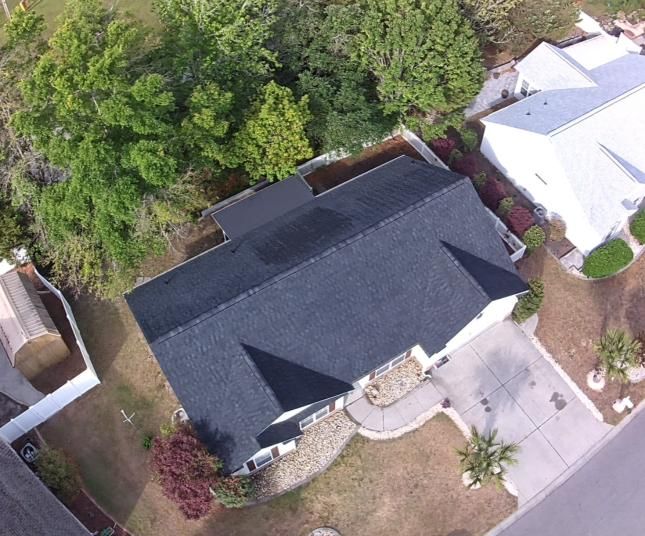 Aerial view of a house with a dark roof, driveway, and surrounding trees in a suburban neighborhood.