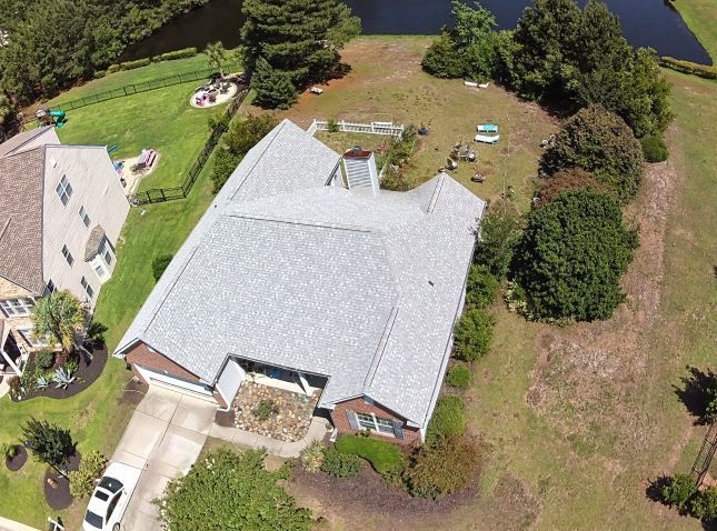 Aerial view of a white-roofed house beside a pond, with a driveway, yard, and trees.