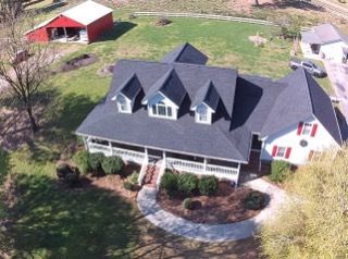 An aerial view of a white two-story house with a black roof, front porch, and red shutters, set on a rural lawn with a barn.