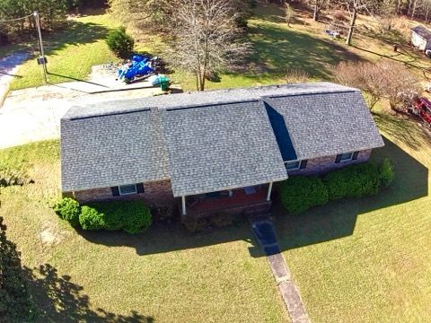 Aerial view of a one-story brick house with a gray shingled roof, surrounded by green lawn and trees.