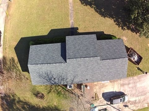 A high-angle aerial view of a house with a dark grey shingled roof, set on a grassy lot with a driveway and parked car.