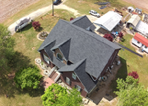 An aerial view of a brick house with a dark shingled roof, surrounded by a yard, driveway, and several smaller buildings.