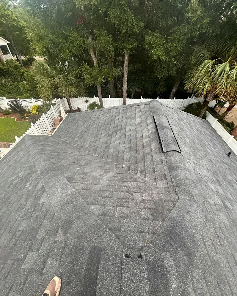 A gray asphalt shingle roof. The roof has some wear. Trees and a fence are in the background.