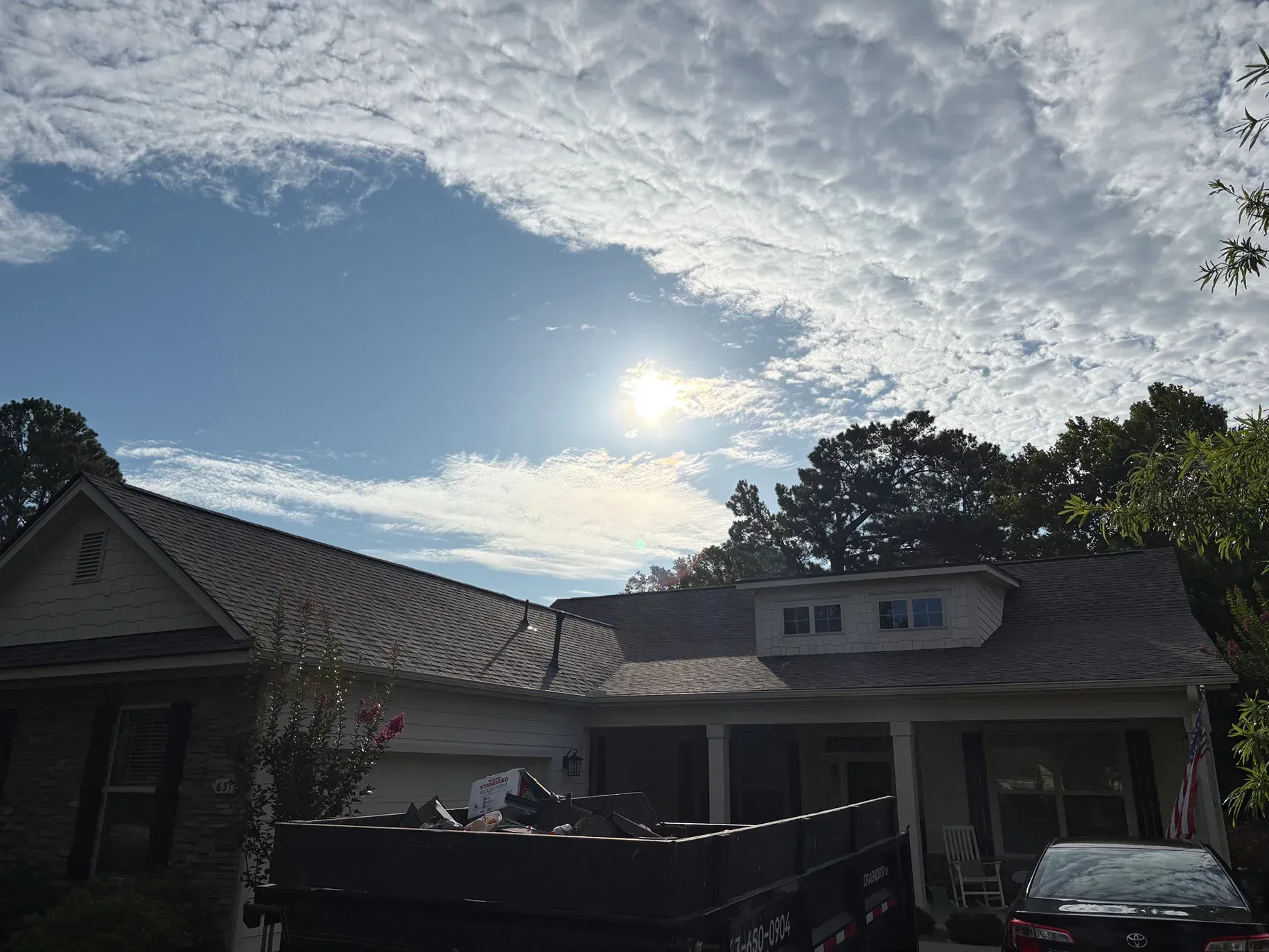House with worn roof, bright sun in cloudy sky, dumpster in front.