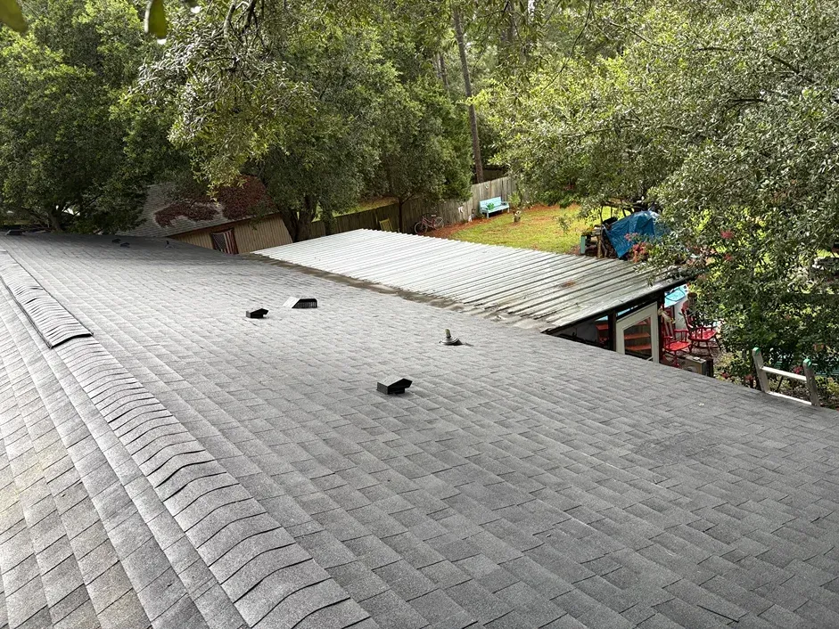 Overhead view of a gray shingled roof with trees in the background. Another lower roof is in the midground.