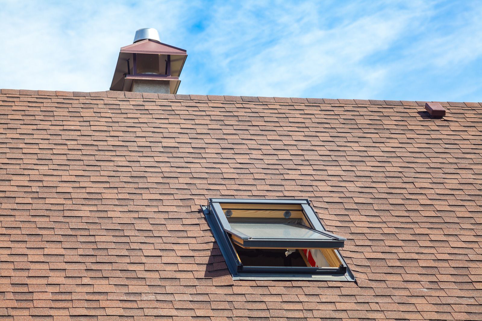 Brown shingled roof with a chimney and open skylight against a blue sky.