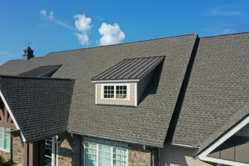 Gray shingle roof with a small metal dormer, under a blue sky with fluffy white clouds.