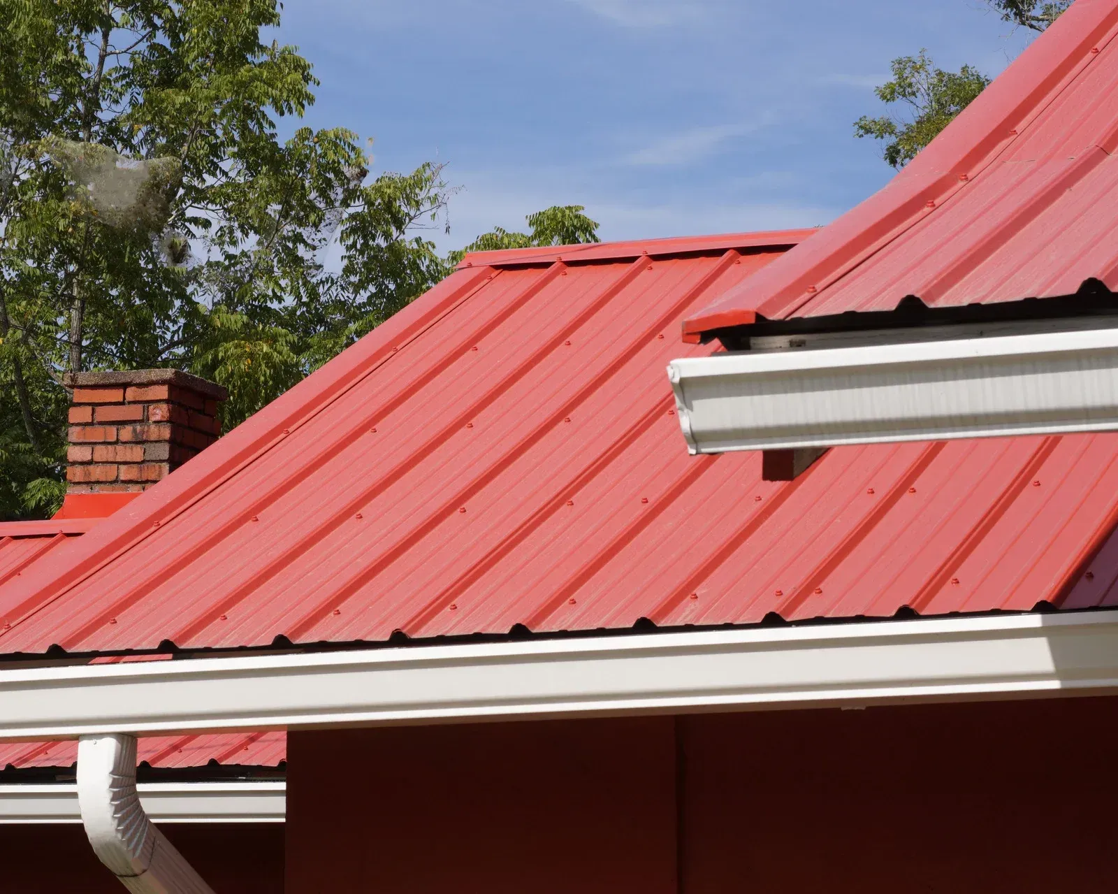 Red metal roof with white gutters and brick chimney against a blue sky.