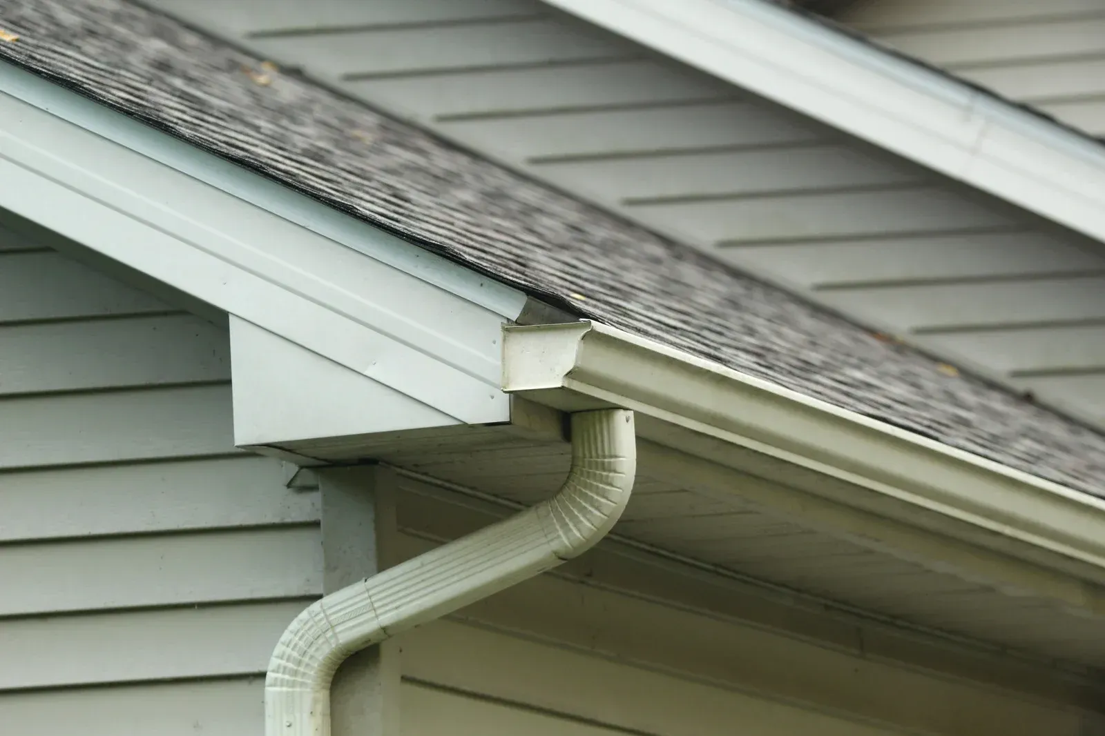 Close-up of a house's roof, gutter, and downspout. The siding is light blue.