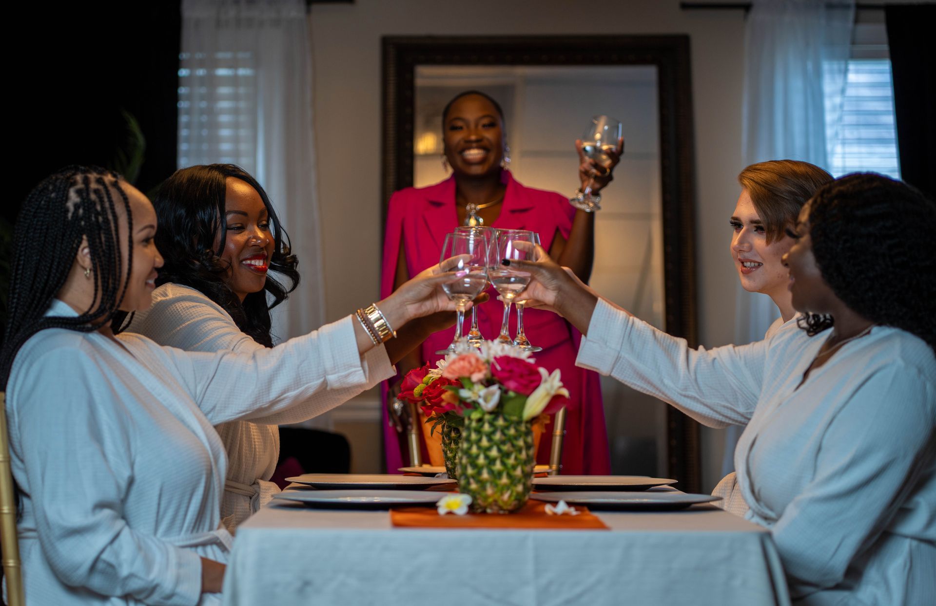 Five women toasting with wine glasses around a table; one woman in pink, others in white robes. Brunch bridal showers