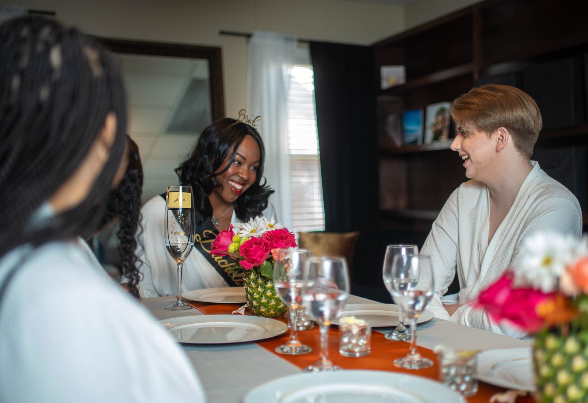 A bride-to-be laughs with a friend at a decorated table, with flowers and glasses.