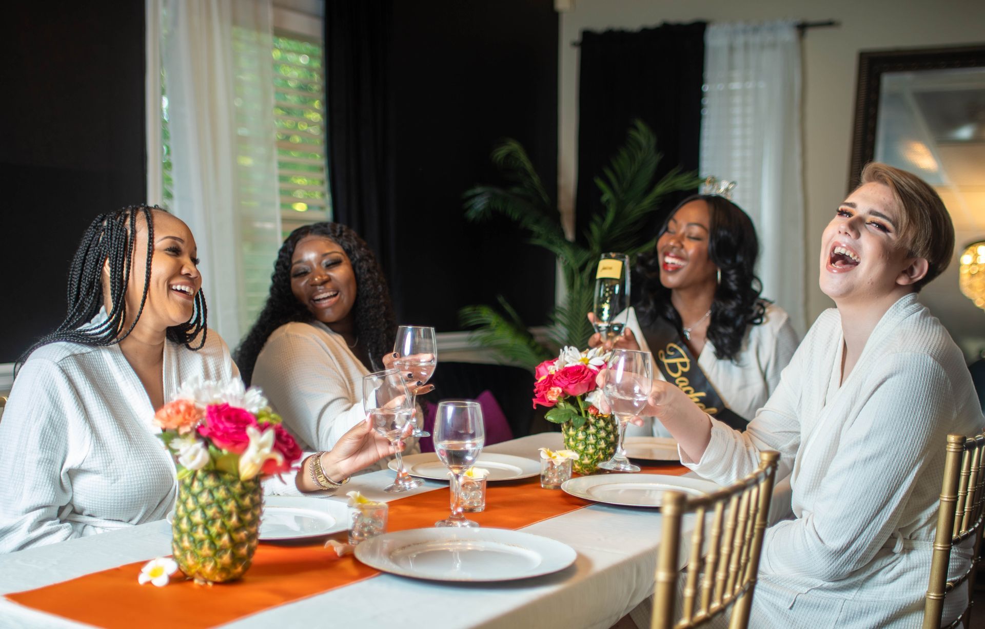 Four women in robes laughing around a table with drinks and flowers.