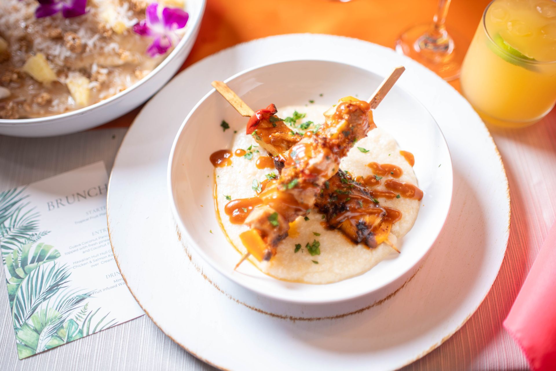 Grilled chicken skewers over grits, drizzled with sauce in a white bowl. Brunch setting.