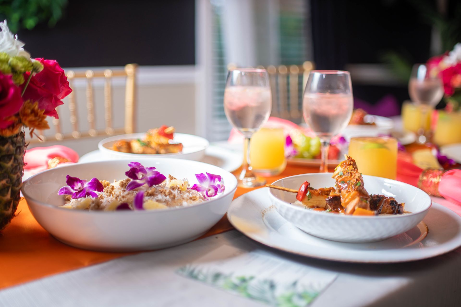 Elegant table setting with food and drinks; white bowls, gold chairs, and colorful floral arrangements.