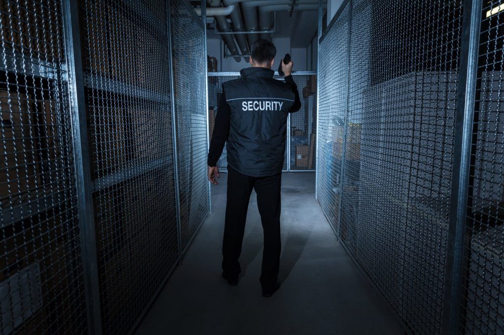 Security guard in black uniform walking down a storage aisle, holding a radio.