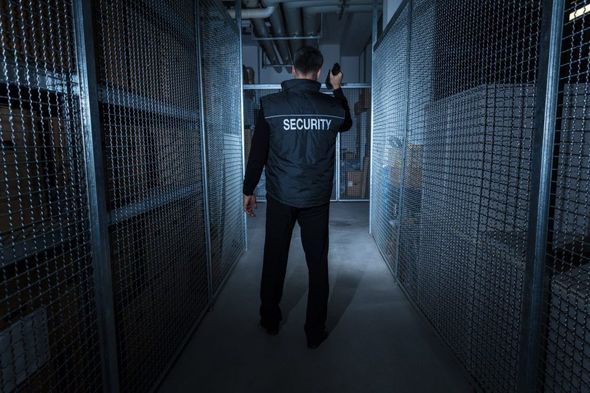 Security guard in black uniform walking down a storage aisle, holding a radio.