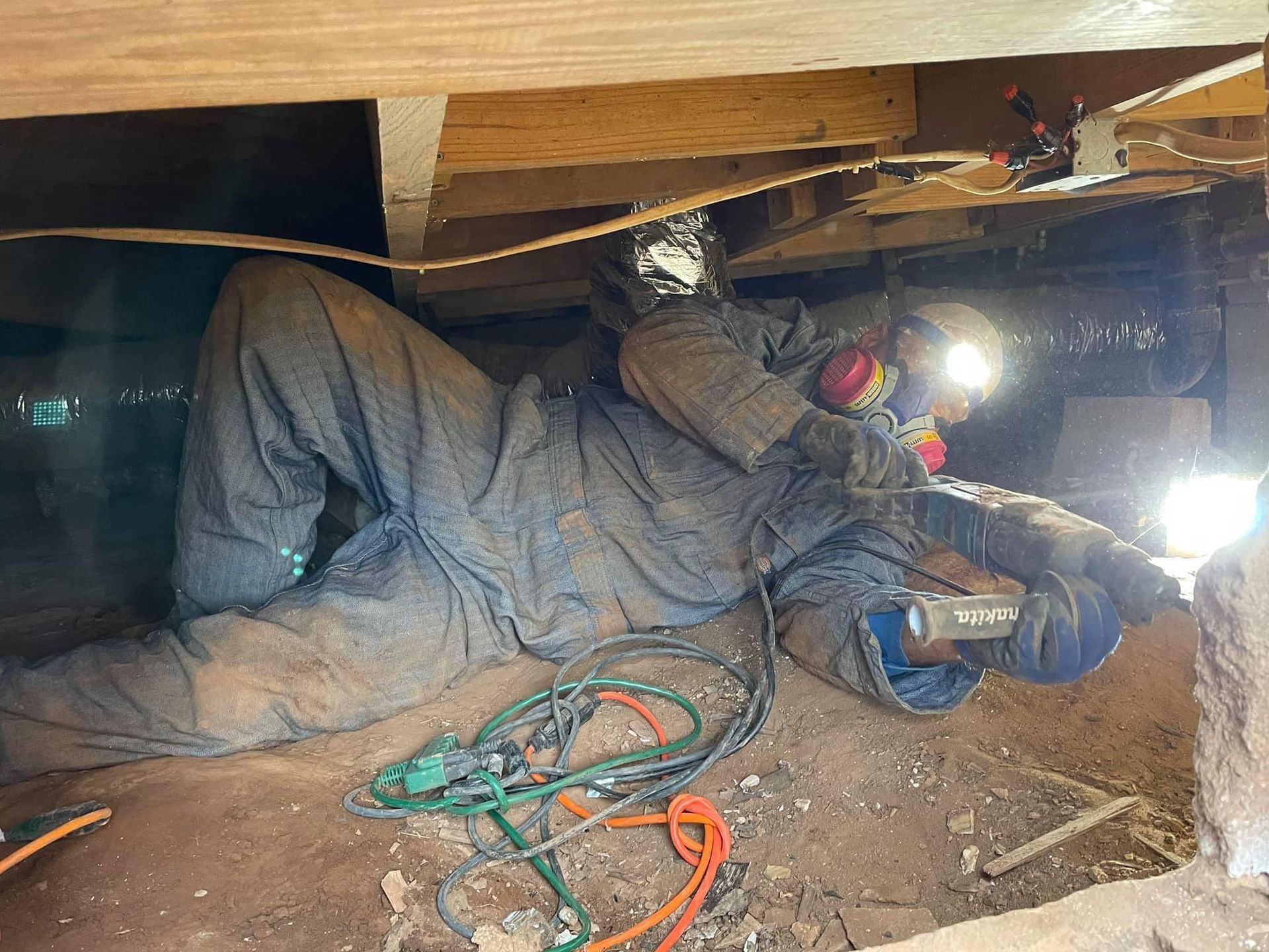 Person wearing protective gear works under a house. They use a light and tool, possibly repairing something.