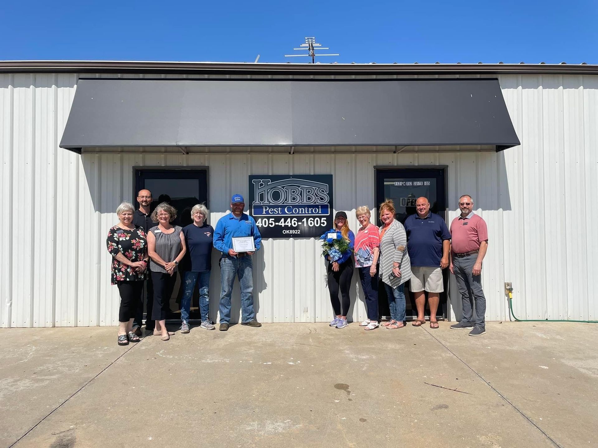 Group of people outside a business holding a certificate and flowers; sunny day.
