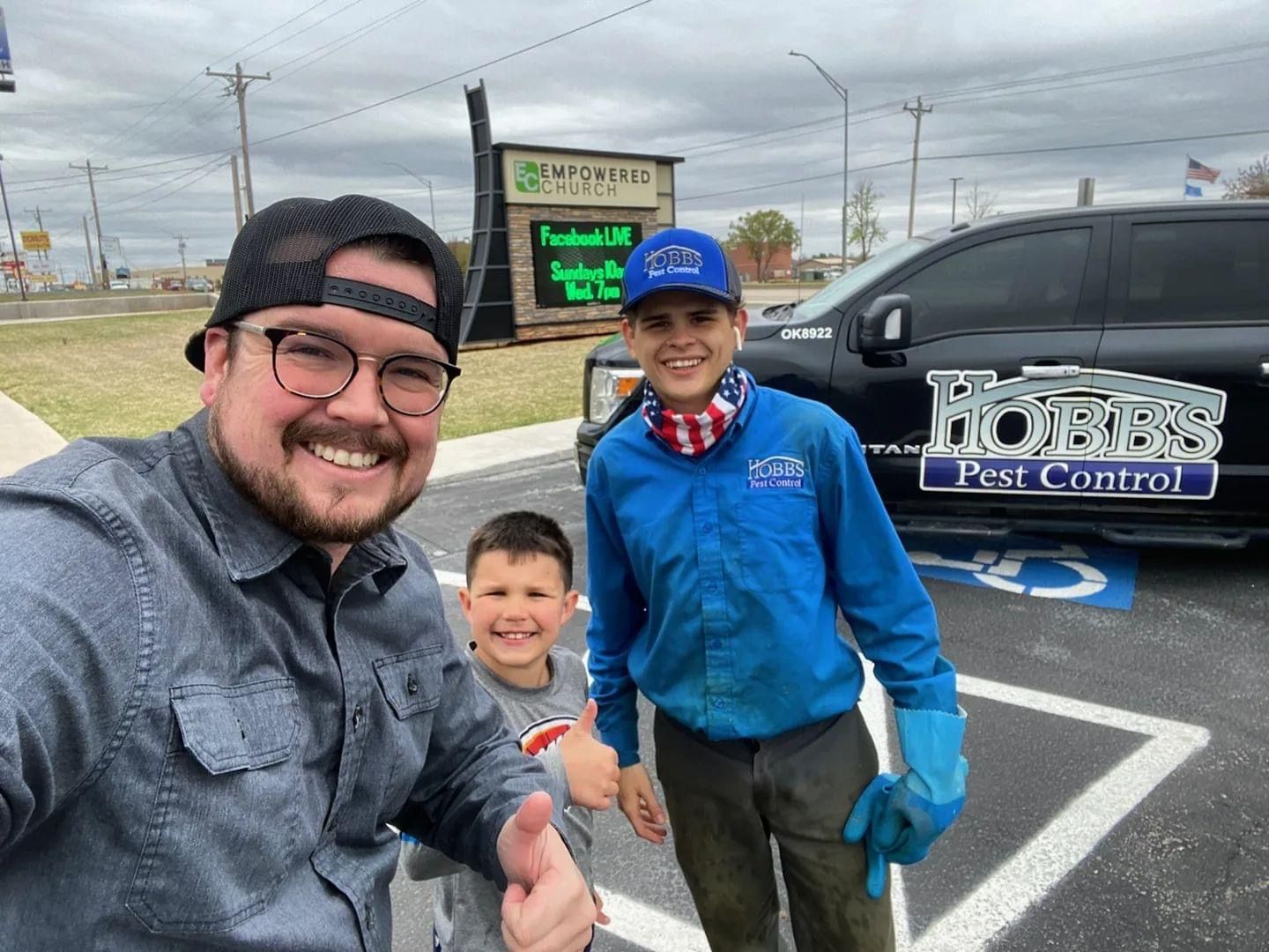 Three people pose for a selfie in front of a pest control business on an overcast day.