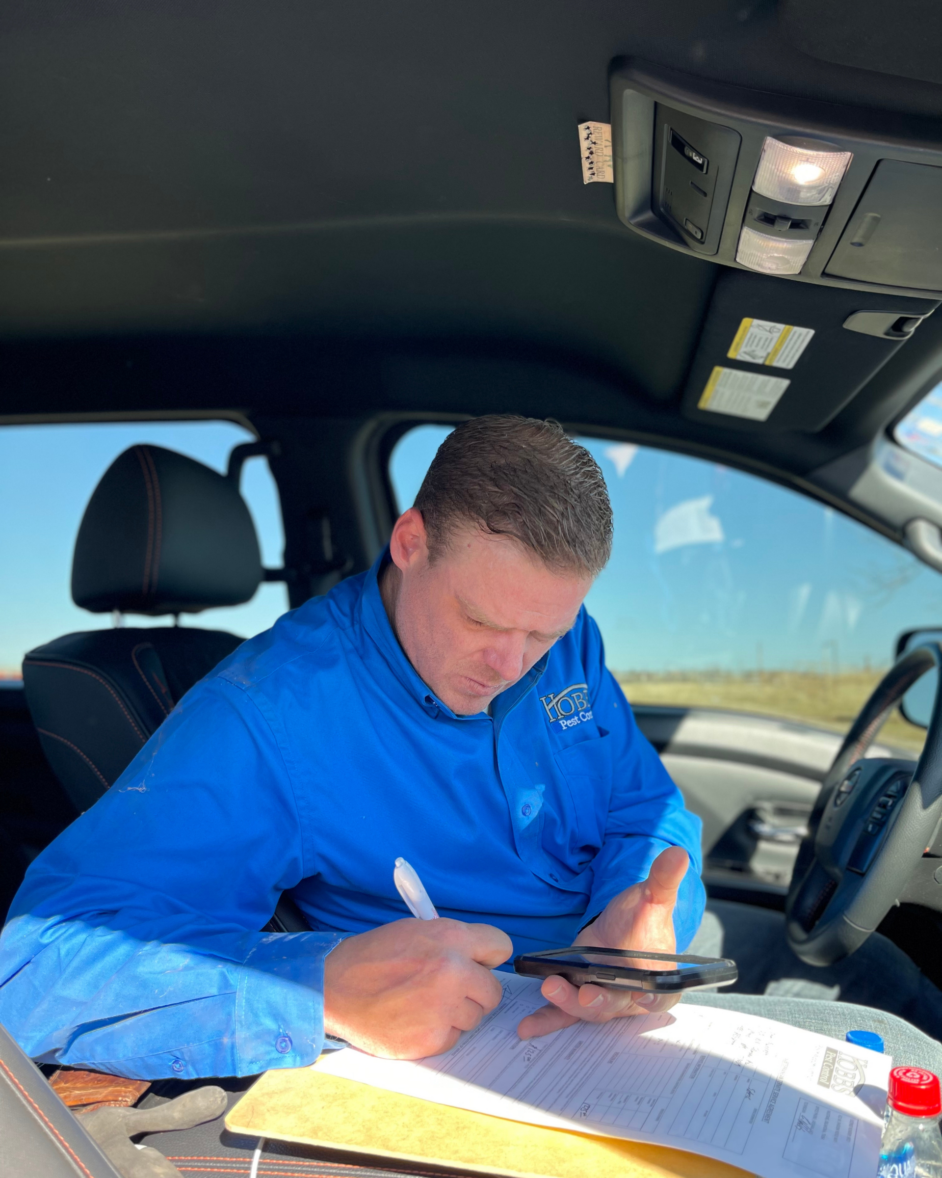 Man in blue shirt inside a vehicle, looking at phone, holding paper and pen. Sunlight streams through window.