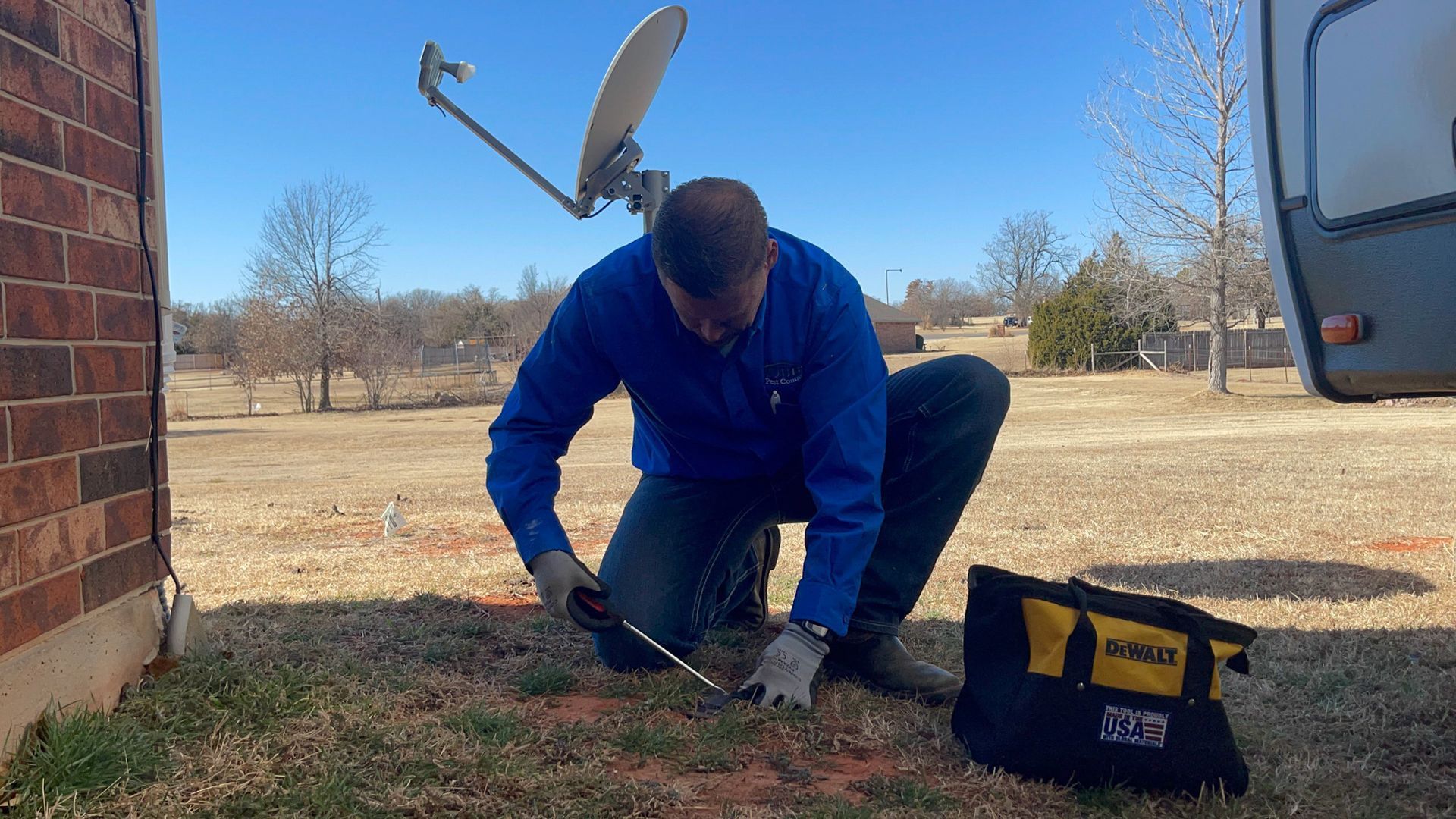 A person kneels near a brick wall, working on the ground with a tool next to a toolbox, with a satellite dish visible.