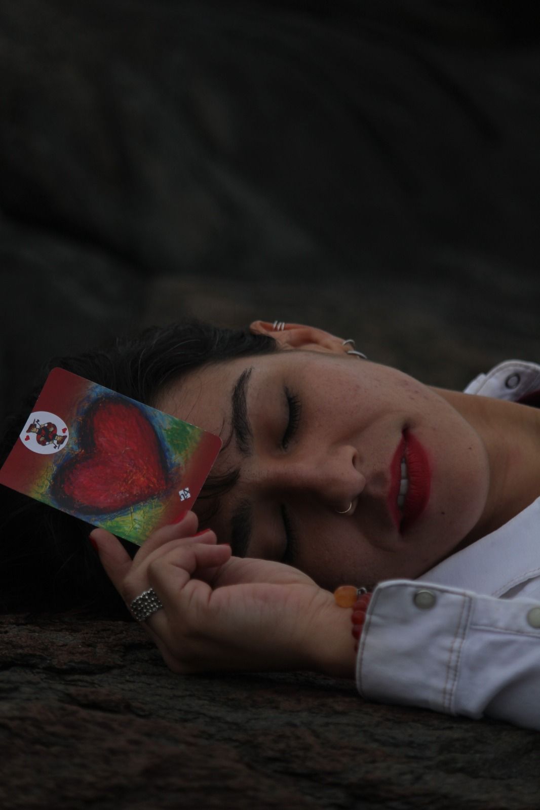 Mulher deitada, segurando um cartão com um coração vermelho, olhos fechados, sorrindo.