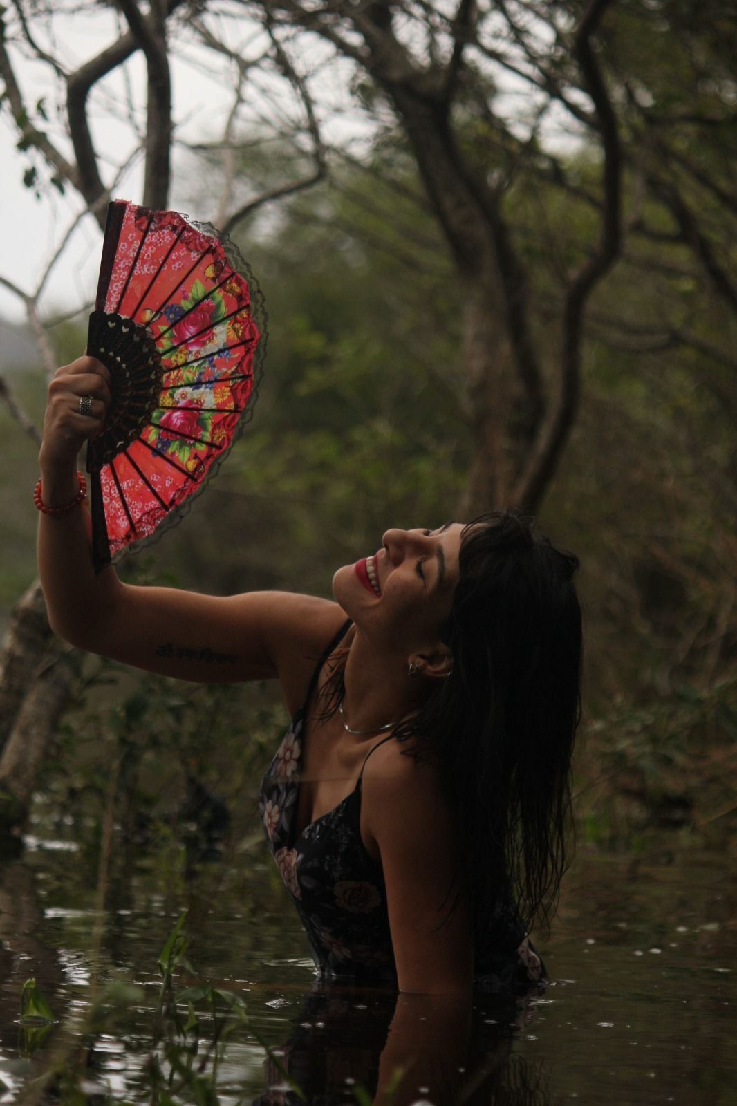 Mulher com vestido floral segurando um leque vermelho, sorrindo em um riacho na floresta.