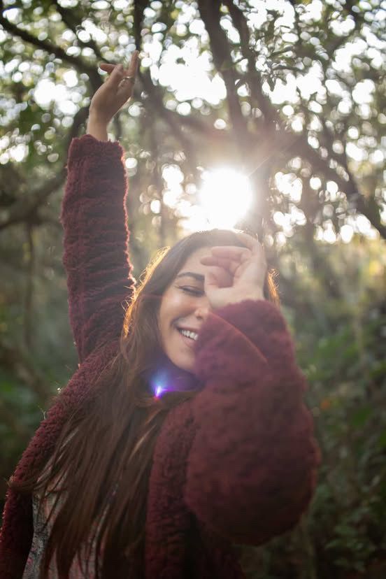 Mulher com longos cabelos castanhos sorri, levanta o braço em direção ao sol que se filtra pelas árvores da floresta; vestindo um suéter vermelho.