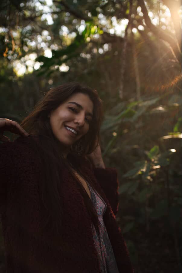 Mulher sorrindo em uma floresta, com a luz do sol filtrando-se pelas árvores. Ela veste um casaco vermelho-escuro e cabelos longos.