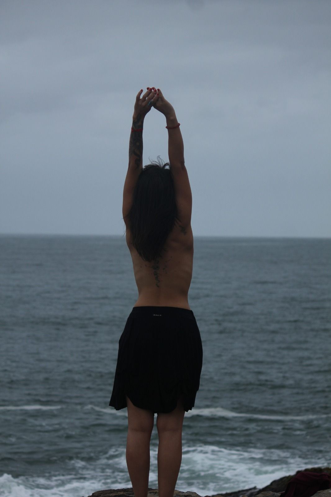 Mulher com os braços levantados, de costas para o observador, em pé em um penhasco com vista para o oceano em um dia nublado.