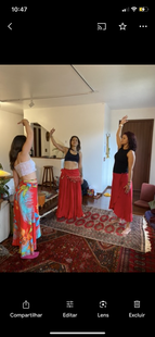 Três mulheres dançam, braços erguidos, vestindo tops curtos e saias. Dentro de casa, sobre um tapete.