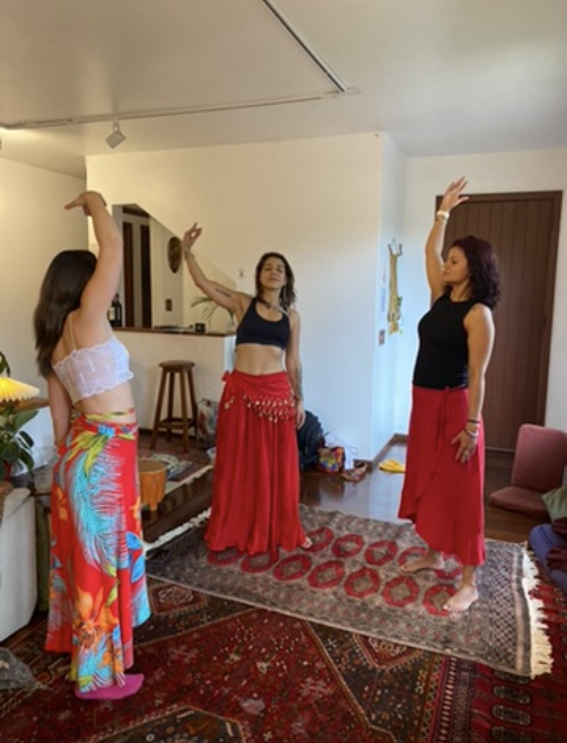 Três mulheres em uma casa praticam dança do ventre, com os braços para cima, usando saias vermelhas.
